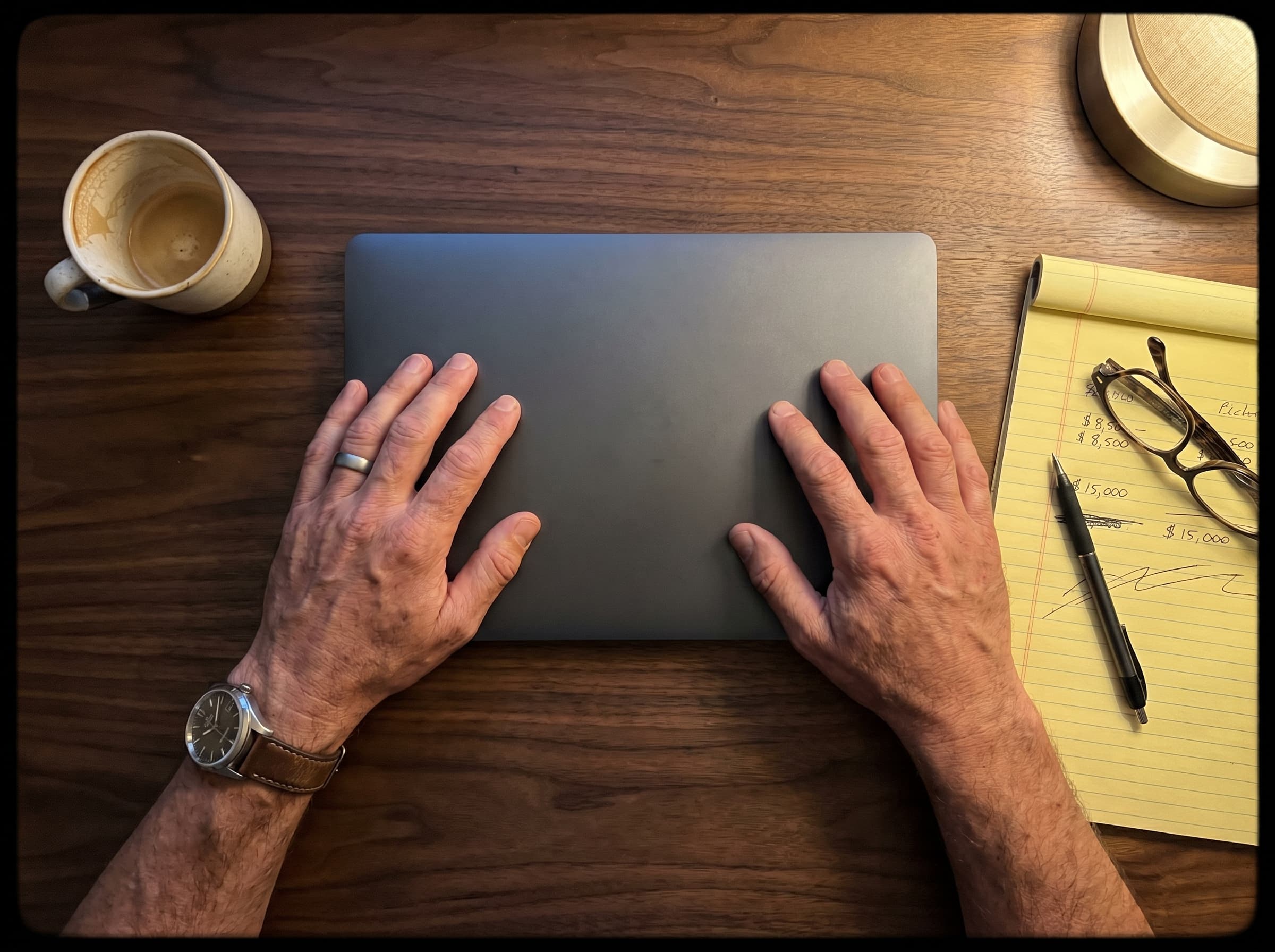 Hands resting on a closed laptop next to a notepad with cost figures